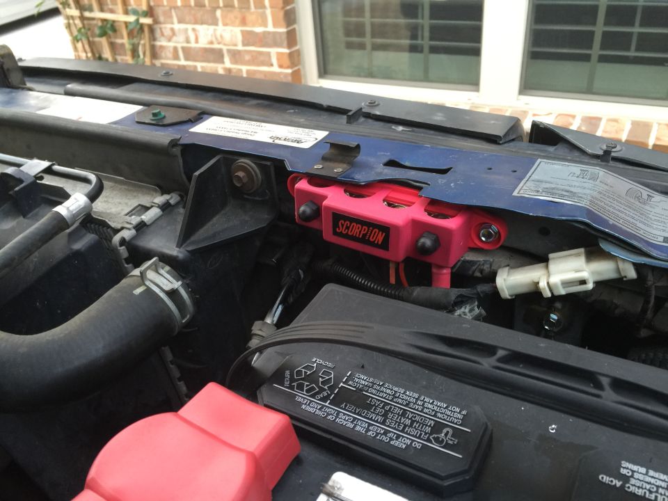 Engine bay with a pink Scorpion relay box mounted on blue metal. A car battery is visible below