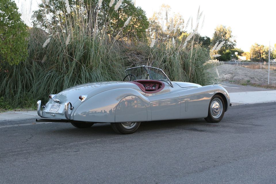 Gray 1952 Jaguar XK120 convertible parked on asphalt with tall grass behind it