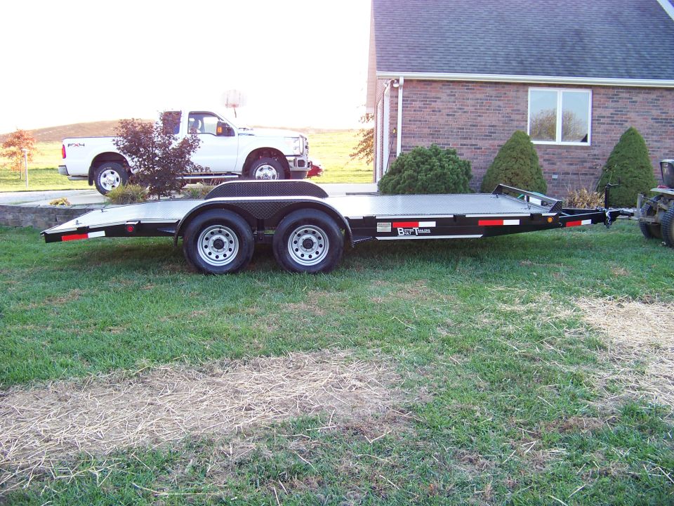 Black car hauler trailer with tandem axles parked on grass, white pickup truck behind it.