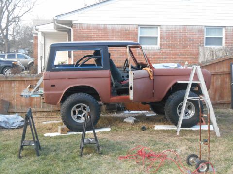 1972 Ford Bronco - A rusty, two-door 1972 Ford Bronco is parked in a grassy yard next to a brick house. The vehicle is missing its hood and appears to be unde…