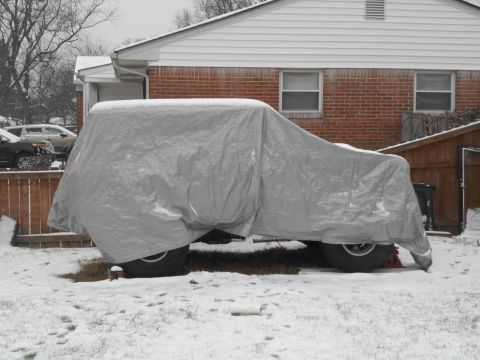 1972 Ford Bronco - A large vehicle, possibly a 1972 Ford Bronco, is covered by a gray tarp and sits in a snowy yard next to a brick house.