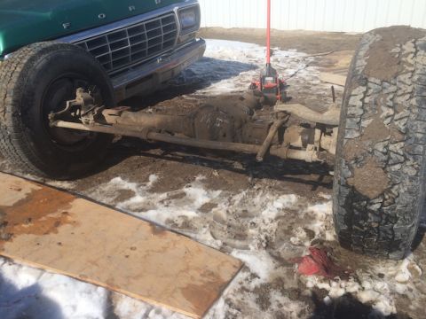 1981 Ford Bronco - The front axle and wheel of a green Ford truck are visible, partially submerged in mud and snow. A red jack is positioned behind the axle,…