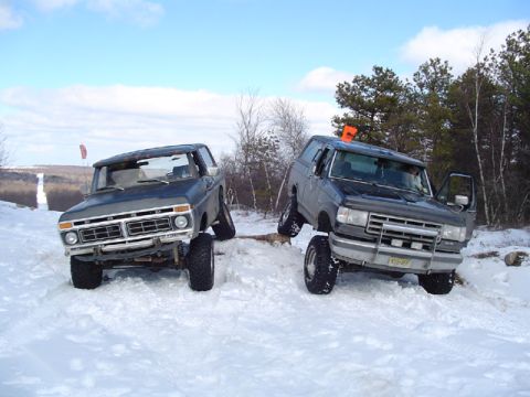 1979 Ford Bronco - Two lifted Ford 4x4 SUVs parked side by side in the snow, both flexed on a mound: a doorless 1970s dark blue Bronco with a chrome front bum…