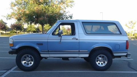 1996 Ford Bronco - The side profile of a two-tone blue and silver Ford Bronco is shown parked in an asphalt lot with trees in the background. The vehicle feat…