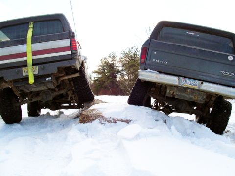 1979 Ford Bronco - Two 1979 Ford Broncos are shown off-roading in a snowy environment, with one vehicle's rear end angled upwards on a rock.