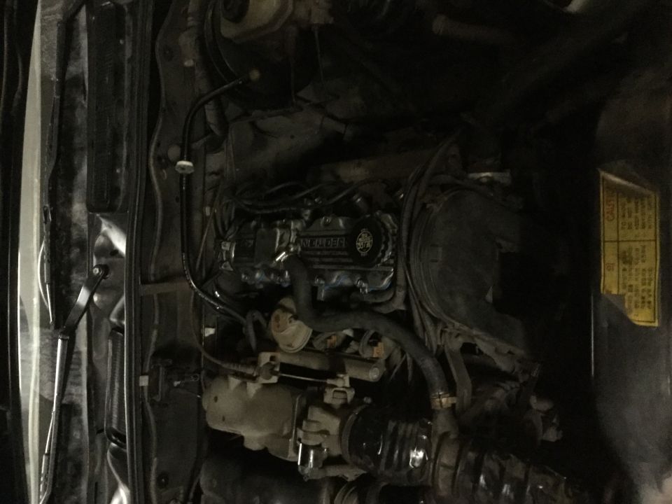 Overhead view of a car engine bay with hoses, wires, and a yellow caution sticker.
