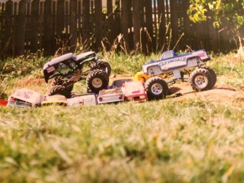 1989 Ford Bronco - Two remote-controlled monster trucks are positioned in a grassy outdoor setting with a wooden fence in the background. One truck, black wit…
