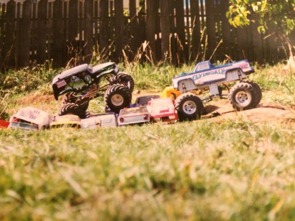 Two RC monster trucks, one black and green, the other white and blue, in a yard.