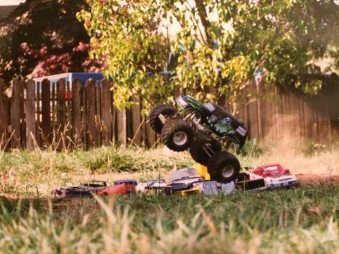 1989 Ford Bronco - A monster truck toy, appearing to be a Grave Digger replica, is airborne over several crushed toy cars. The scene is set outdoors with gras…