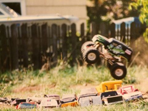 1989 Ford Bronco - A monster truck toy is captured mid-air, jumping over a line of toy cars and a yellow toy dump truck in a grassy yard.