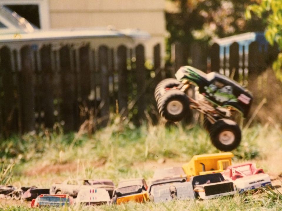 Toy monster truck jumping over toy cars and a dump truck in a grassy yard.