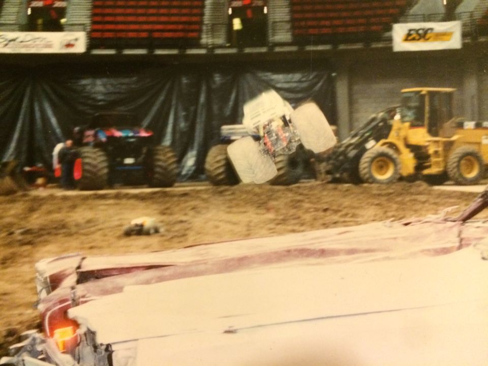 Monster truck in mid-air, loader vehicle, and another monster truck in background at an indoor event