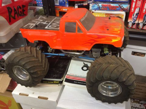 1989 Ford Bronco - A bright orange RC monster truck with flame decals on the hood sits on top of stacked boxes. The truck has large, knobby tires and a red bo…