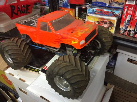 1989 Ford Bronco - An orange RC monster truck with flame decals on the hood sits on top of stacked white boxes. The truck has large, knobby tires and visible…