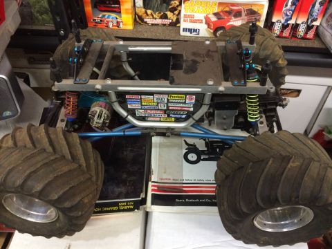 1989 Ford Bronco - A custom-built RC monster truck chassis with large, treaded tires is shown in a close-up, angled view. The chassis features a silver metal…