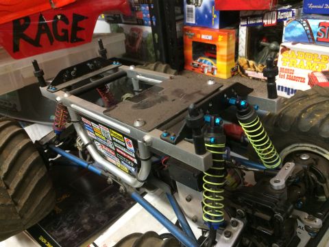 1989 Ford Bronco - A close-up, low-angle shot of the chassis and suspension of a remote-controlled monster truck with a red body labeled "RAGE". The truck fea…
