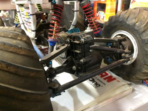 1989 Ford Bronco - Close-up view of the rear suspension and drivetrain of a monster truck, featuring large tires, red-sprung shocks, and a black gearbox.