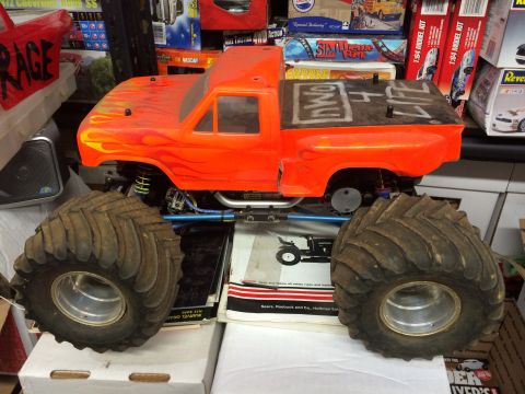 1989 Ford Bronco - An orange remote-controlled monster truck with flame graphics is shown from a 3/4 front angle, resting on top of boxes and papers. The truc…