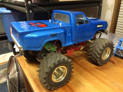 1989 Ford Bronco - A blue remote-controlled monster truck with large tires and a red decal on the bed sits on a wooden surface. The truck has visible suspensi…