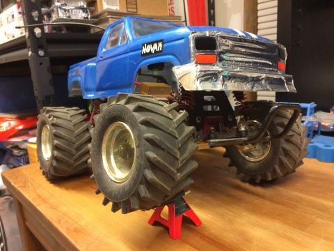 1989 Ford Bronco - A blue RC monster truck with oversized tires is displayed on a wooden surface, supported by red jack stands. The front grille appears to be…