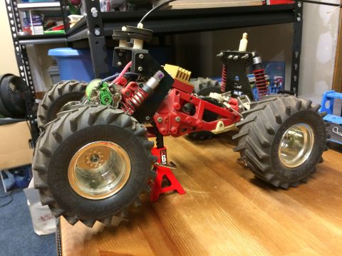 1989 Ford Bronco - A custom-built remote-controlled monster truck with large knobby tires and a red chassis is displayed on a wooden surface, supported by a r…
