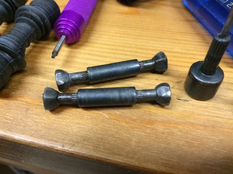 1989 Ford Bronco - Two black metal drive shafts with splined ends and flared ends are laid out on a wooden surface. A purple handled tool and other black part…