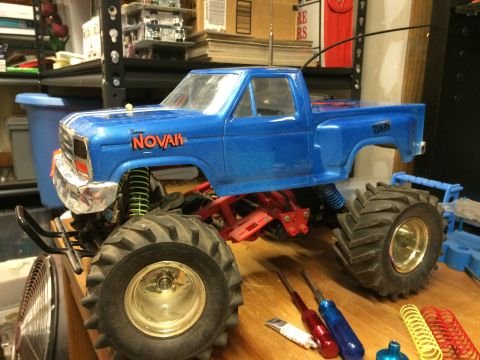1989 Ford Bronco - A blue, modified remote-controlled monster truck with large tires and chrome accents sits on a wooden surface. Visible text on the truck in…