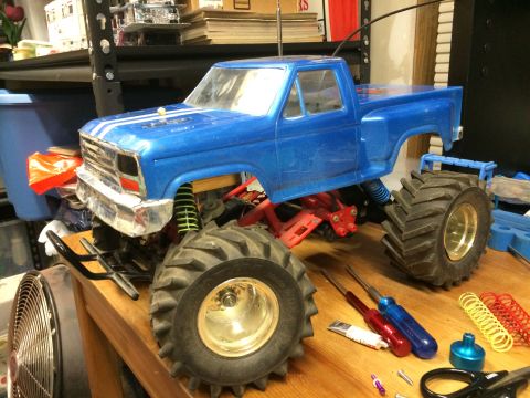 1989 Ford Bronco - A blue RC monster truck with large tires and chrome accents is on a wooden surface with tools and spare parts.