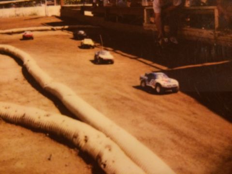 1989 Ford Bronco - Several remote-controlled cars race on a dirt track with large, beige foam barriers lining the course. One car in the foreground is a white…