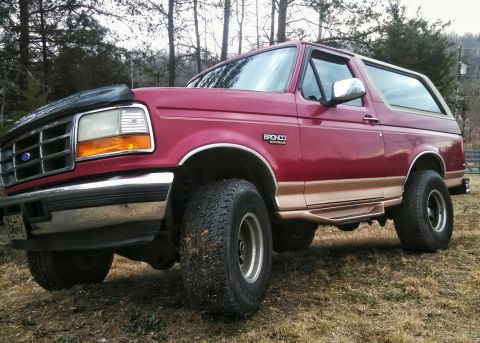 1995 Ford Bronco - This red Ford Bronco is shown with a lift kit and large tires, parked on a grassy area with trees in the background.