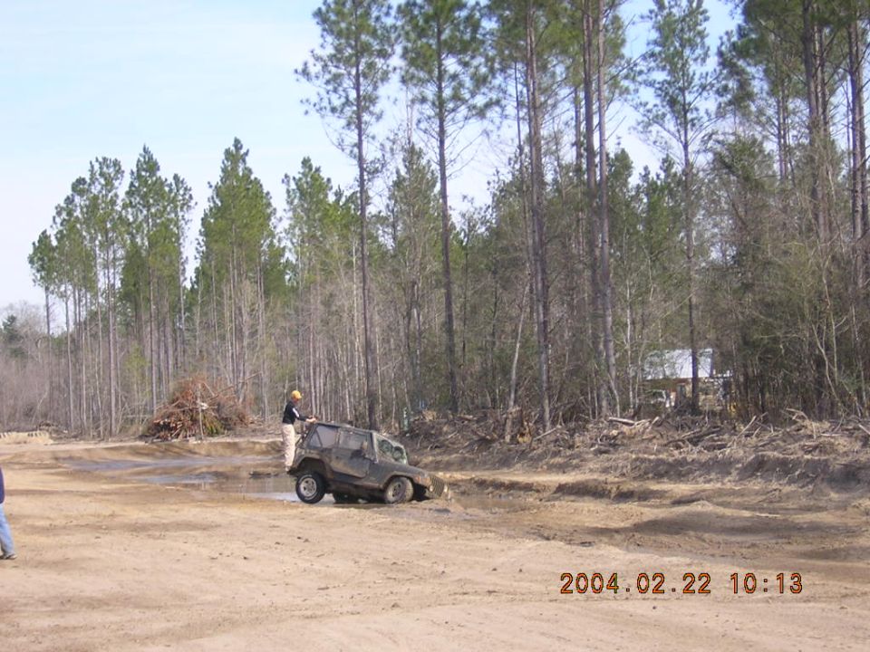 Off-road vehicle stuck in mud with a person standing nearby. Trees and debris in the background
