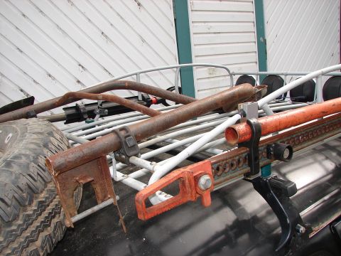 1964 Volkswagen Beetle - Hi-lift jack and spare tire on a rack, with rusty metal bars and a white building in the background.
