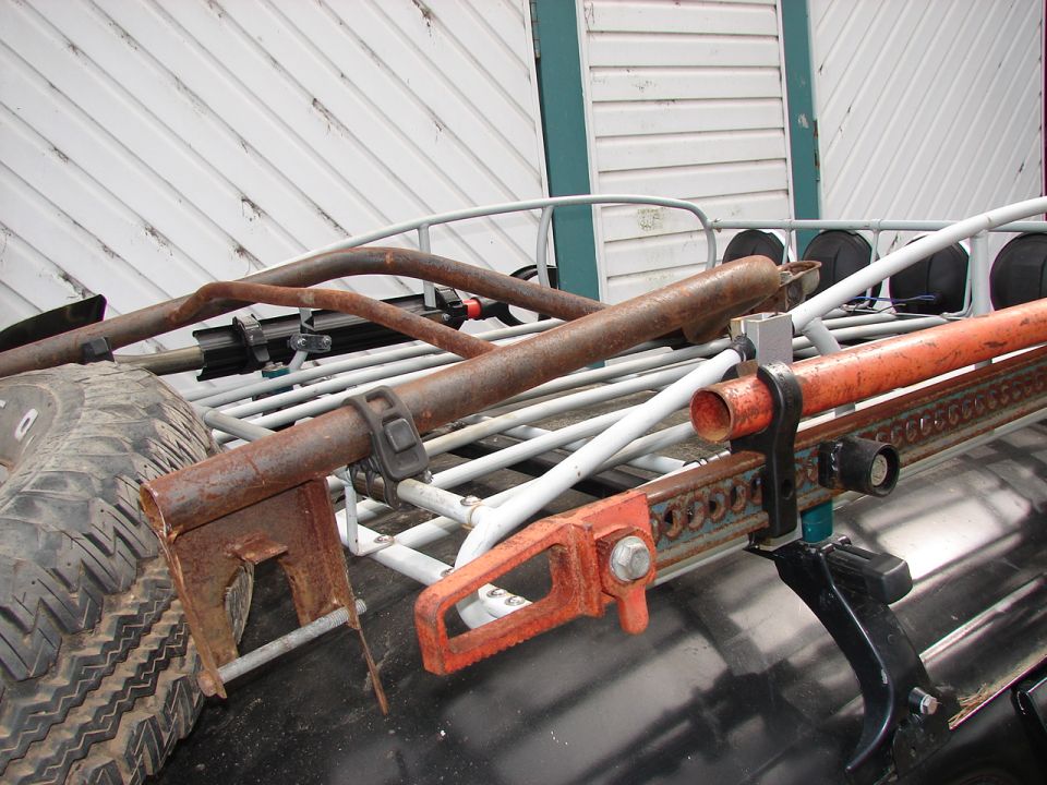 Hi-lift jack and spare tire secured to a roof rack. Some rusty metal bars are also visible