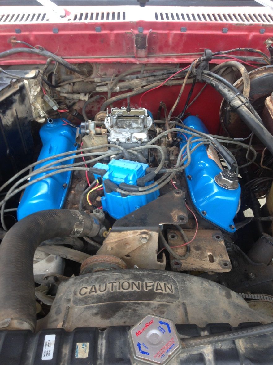 Engine bay with blue valve covers and a carburetor. Radiator fan shroud has a caution label