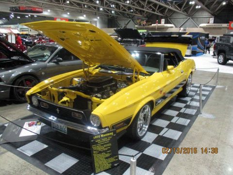 2004 Ford Crown Victoria - A bright yellow 1972 Ford Mustang convertible is displayed with its hood open, revealing the engine bay. The car is on a checkered display…