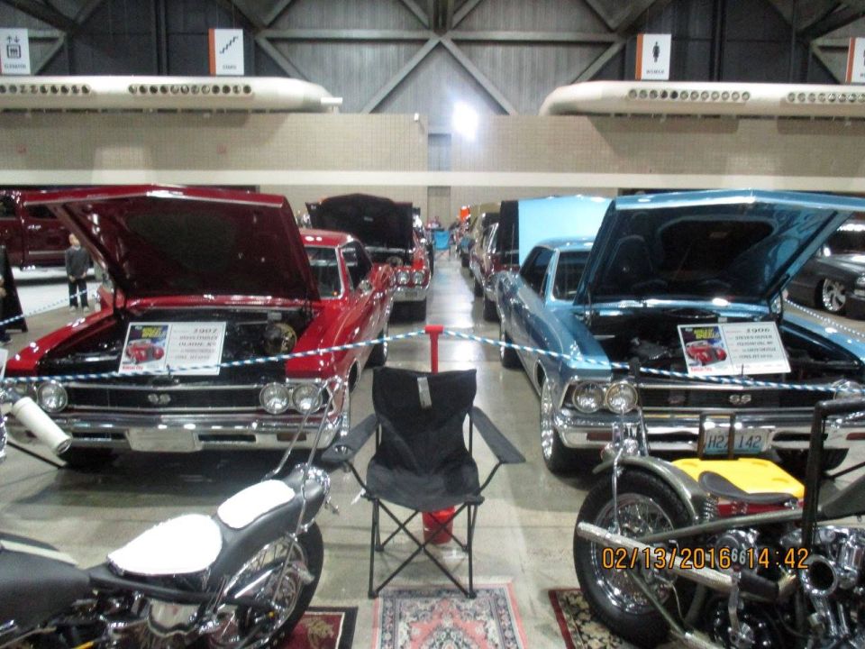 Two classic cars, a red and a blue one, with hoods open, parked indoors with motorcycles in front
