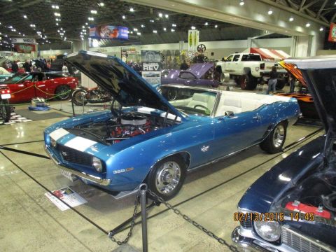 2004 Ford Crown Victoria - A blue 1969 Chevrolet Camaro SS convertible is displayed with its hood open, showing the engine. Other vehicles and displays are visible in…