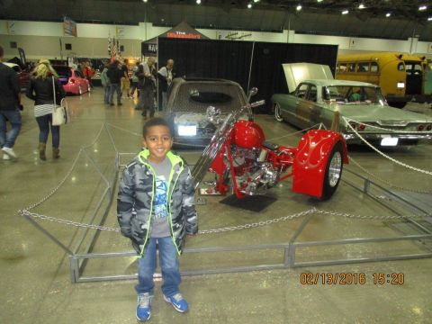 2004 Ford Crown Victoria - A young boy poses in front of a red trike motorcycle and several vintage cars at an indoor car show.