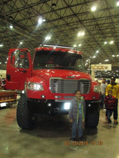 2004 Ford Crown Victoria - A red truck with a prominent light bar and large tires is displayed indoors, with several people nearby.