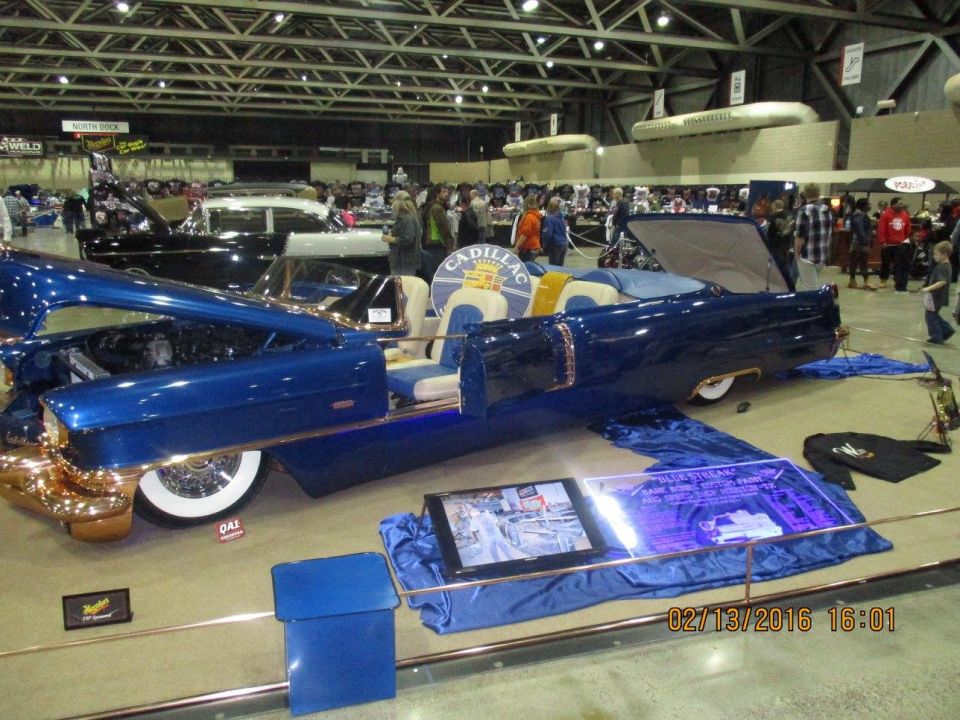 Blue convertible with gold trim and white interior, hood open, on display at a car show