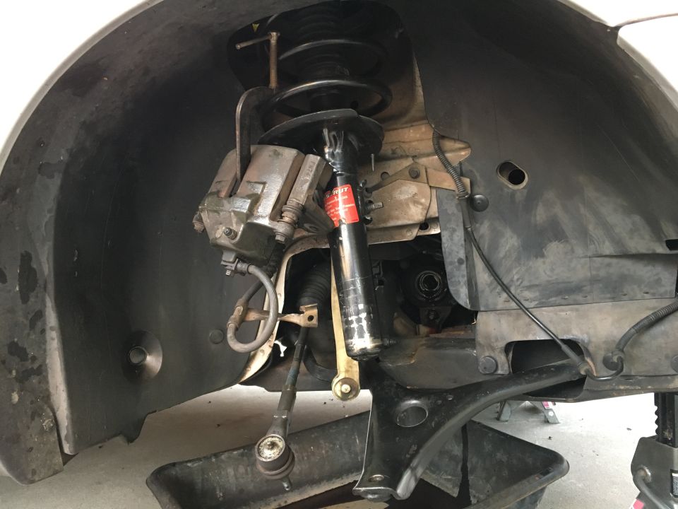 Close-up of car's front suspension with brake caliper, strut, and tie rod end visible. Oil drain pan underneath