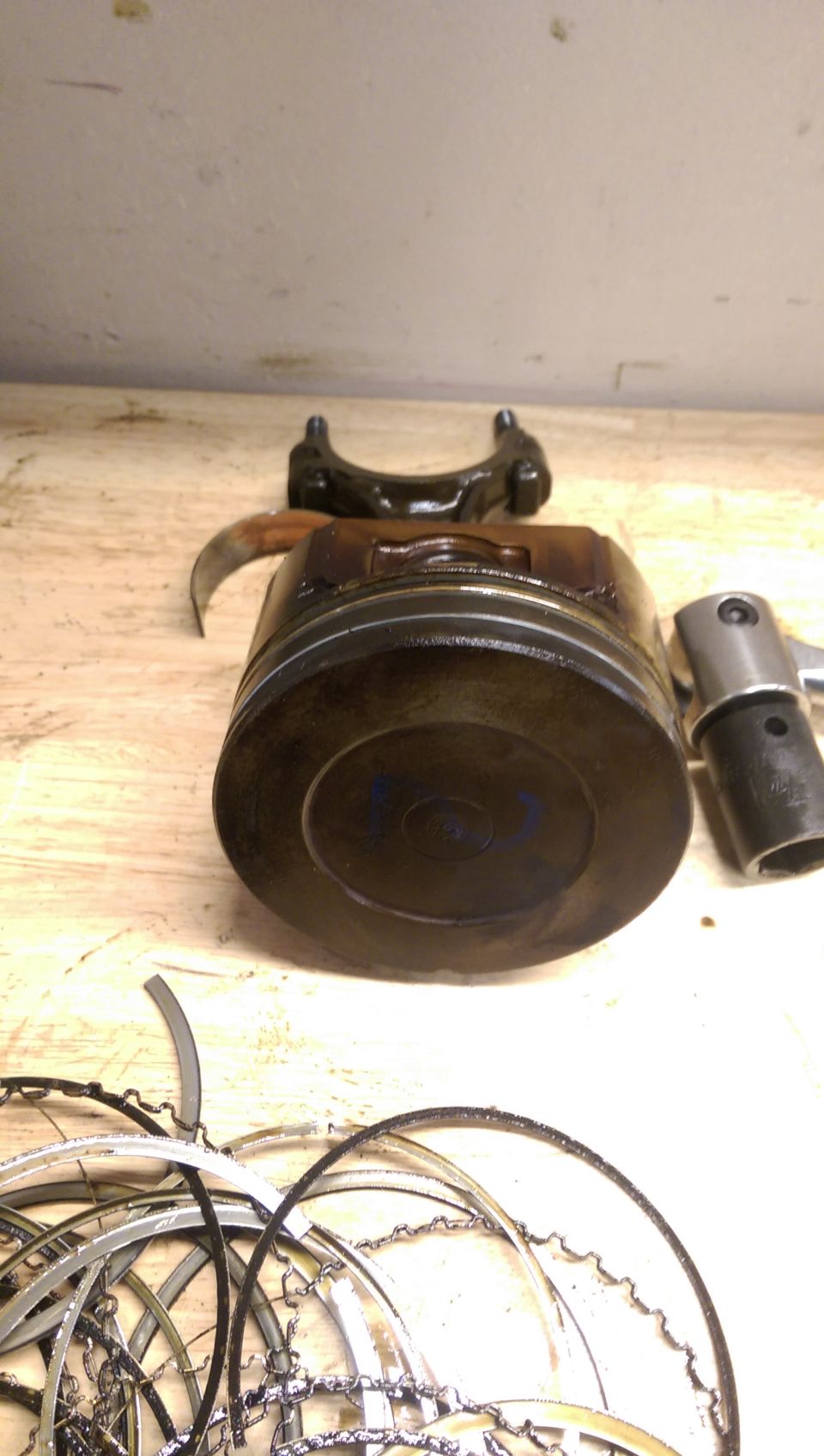Piston with connecting rod attached, plus a pile of piston rings and a tool on a wooden surface