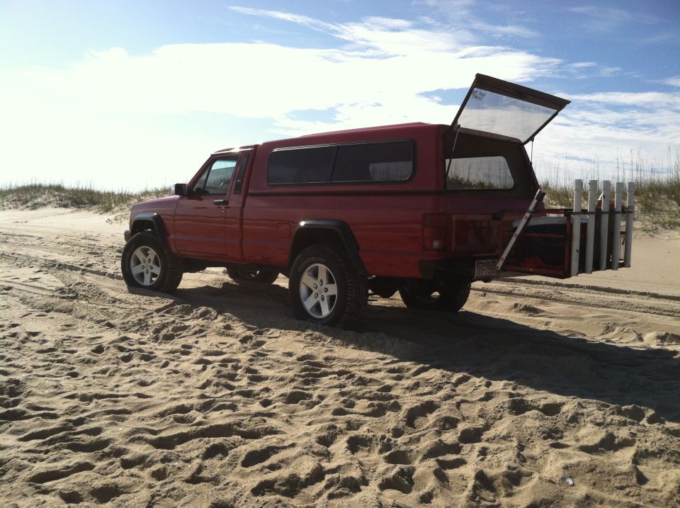 1991 Jeep Comanche pickup with camper shell and fishing rod holders on the tailgate, parked on sand dunes