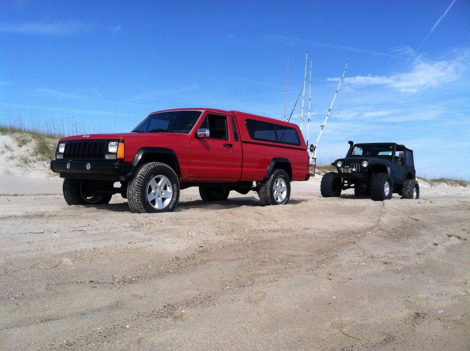 1991 Jeep Comanche pickup with a camper shell and a black Jeep Wrangler parked on a beach with fishing rods in the back of the Comanche