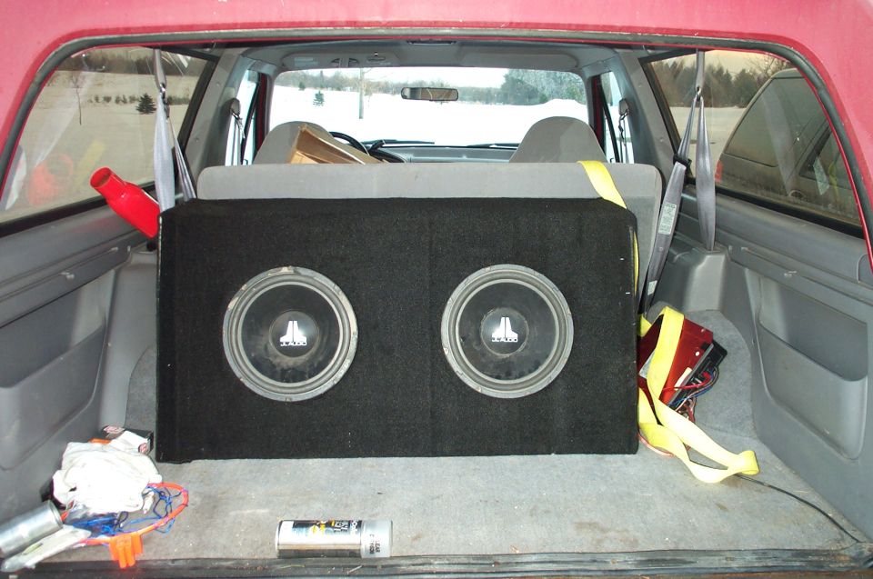 Two JL Audio subwoofers in carpeted box strapped in the rear cargo area of a red SUV