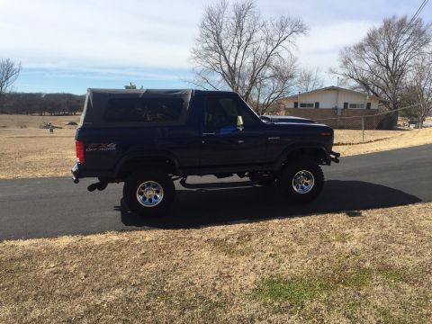 1982 Ford Bronco - A lifted, dark blue Ford Bronco with a soft top is parked on a paved road in a suburban area.