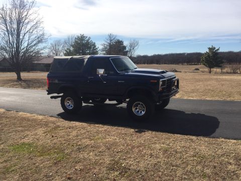 1982 Ford Bronco - This lifted 1982 Ford Bronco features a dark blue exterior and a removable soft top, seen driving on a paved road.
