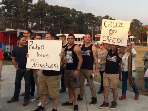1995 Mercury Grand Marquis - Group of men outdoors holding handmade signs reading 'Rubio listens to Nickelback!' and 'Cruz is Creed!' near food trucks.