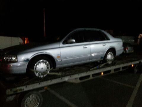 1999 Ford Fairlane - A silver sedan is parked on a flatbed tow truck at night. The vehicle is viewed from the side, showing its front wheel and fender.