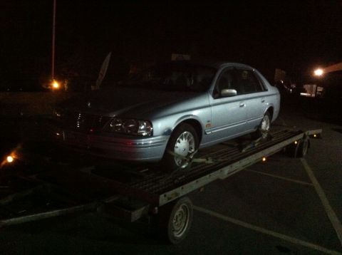 1999 Ford Fairlane - A silver sedan is loaded onto a car trailer at night. The vehicle is strapped down and the trailer has visible lights.